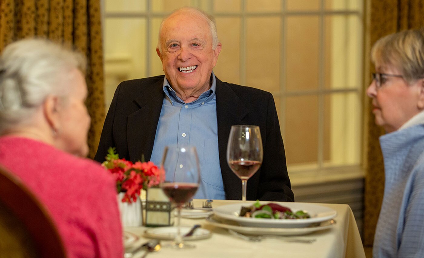 Three residents sit at a dining table. A man looks at the camera smiling with a glass of wine.