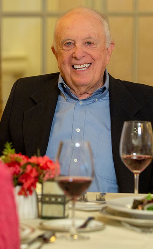 Three residents sit at a dining table. A man looks at the camera smiling with a glass of wine.