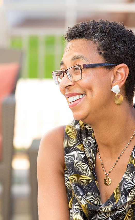 A woman sitting outdoors in the sun with patio furniture in the background.