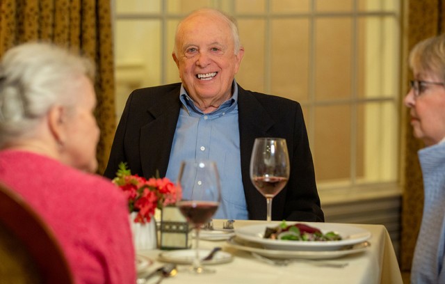 Three residents sit at a dining table. A man looks at the camera smiling with a glass of wine.
