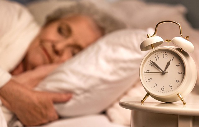 An alarm clock on a nightstand with a woman slipping in the background.