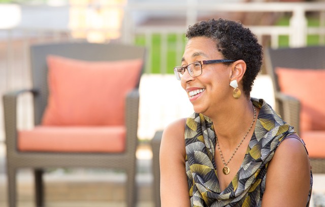 A woman sitting outdoors in the sun with patio furniture in the background.