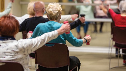 Residents in an exercise class sitting in chair lifting waits facing a mirror.