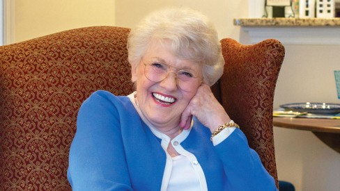 Resident sitting in a chair with her face leaning on her hand smiling.