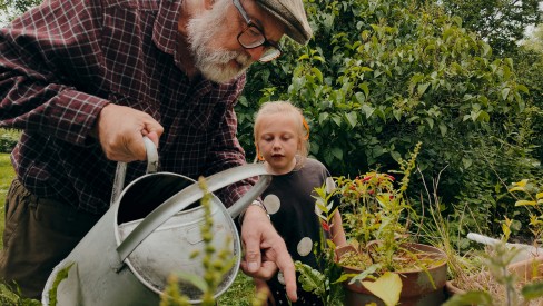 A man waters plants with his grandchild, demonstrating a healthy intergenerational relationship.