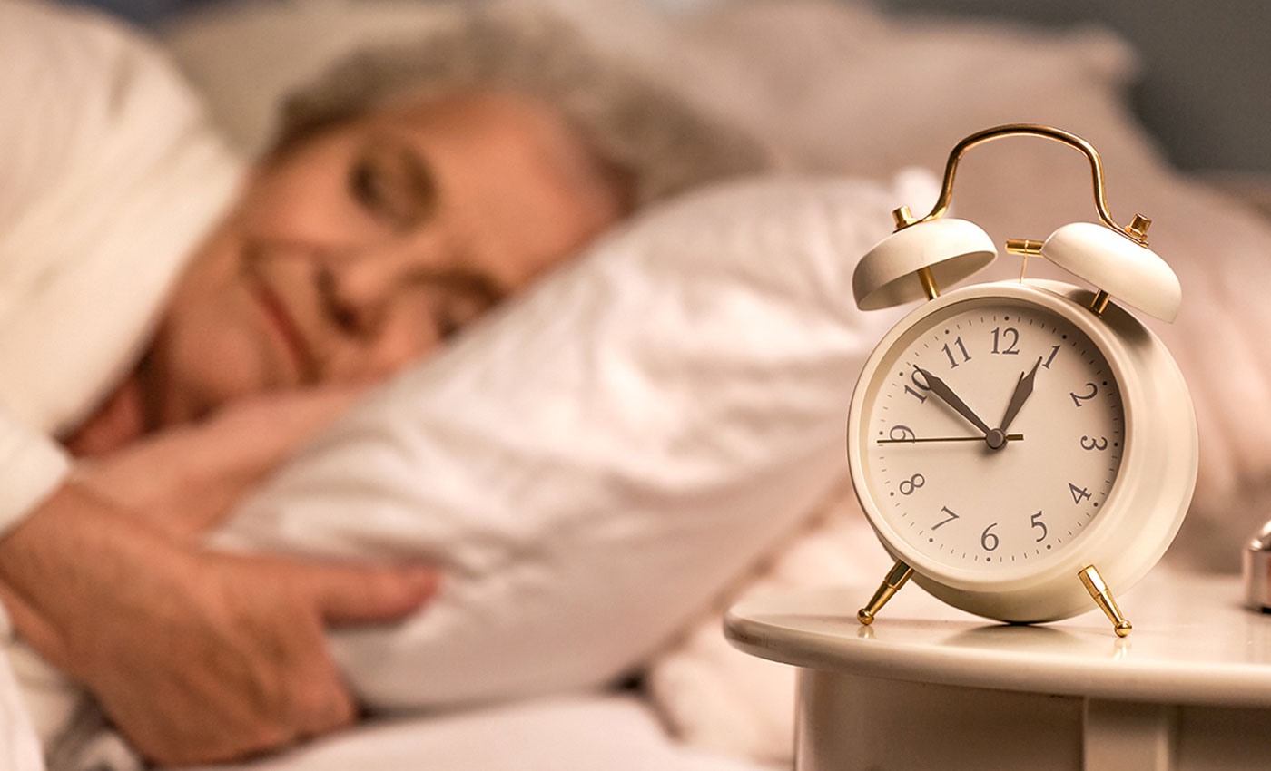 An alarm clock on a nightstand with a woman slipping in the background.