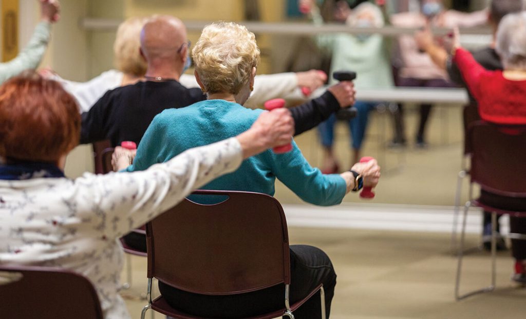 Residents in an exercise class sitting in chair lifting waits facing a mirror.