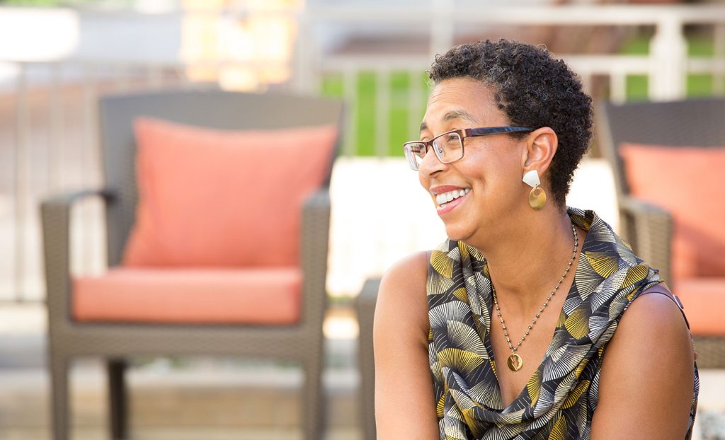 A woman sitting outdoors in the sun with patio furniture in the background.